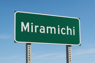 Green road sign with "Miramichi" in white letters, standing beneath a clear blue sky, marking the town's entrance.