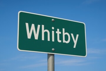 Green road sign displaying "Whitby" beneath a bright blue sky, indicating city entrance, travel route, or Ontario location.