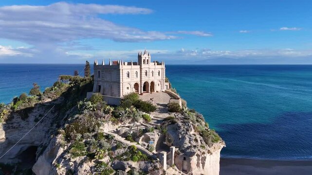 A historic church perched  of Santuario di Santa Maria dell'Isola di Tropeaon a cliff overlooking the ocean, surrounded by lush greenery and a clear blue sky.