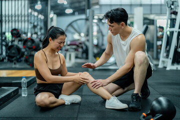 Young woman suffering knee pain during workout at gym, personal trainer assisting