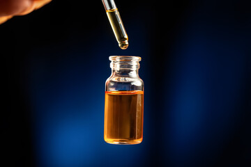 Amber liquid being dispensed from a glass dropper into a small vial against a dark blue background.