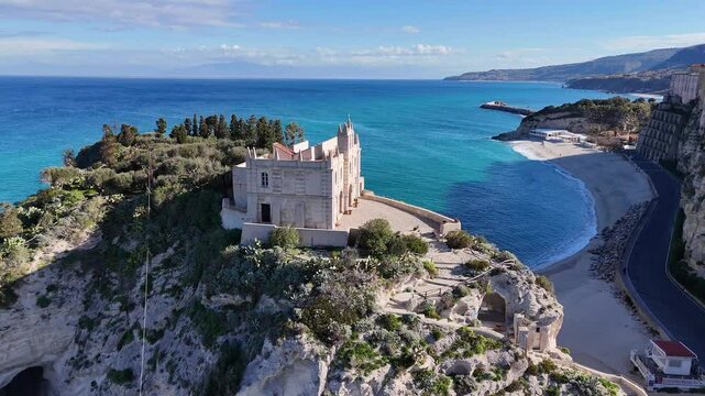 A historic church perched  of Santuario di Santa Maria dell'Isola di Tropeaon a cliff overlooking the ocean, surrounded by lush greenery and a clear blue sky.