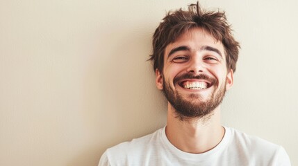 Obraz premium A young man with a beard, wearing a white t-shirt, standing against a plain beige wall with a neutral expression on his face.