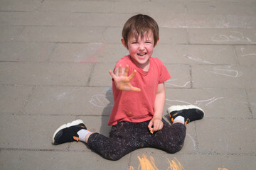 Happy child sitting on concrete floor, extending hand in playful gesture while drawing with sidewalk chalk. Outdoor play and childhood fun concept. A child aged 3 years