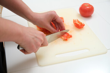 Hands preparing food by chopping a juicy red tomato on a plastic board with a sharp knife in a home kitchen setting.