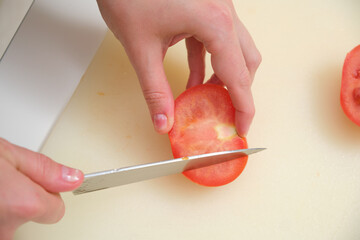 Person's hands slicing a red, juicy tomato with a kitchen knife on a light plastic cutting board during meal preparation.