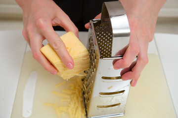 Close-up of hands grating cheddar cheese using a stainless steel box grater on a white cutting board in a kitchen.