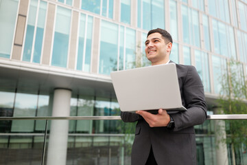 Fototapeta premium Business professional stands confidently outdoors with laptop overlooking modern architecture in a vibrant urban setting
