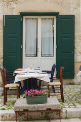 Matera, Italy. Outdoor restaurant tables set for dining in front of a charming old door with green shutters, along a cobbled street in the historic Sassi di Matera, Italy. 