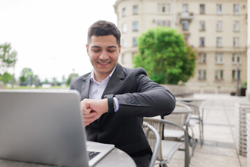 Focused professional checks his smartwatch while working on his laptop outdoors in a charming urban...