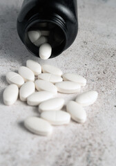 White oval pills are scattered from an overturned black bottle onto a textured grey surface,...