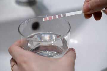 Hand testing clear tap water quality with a color-changing pH strip in a glass for home analysis,...