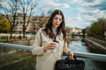 Young caucasian woman walk home from work drink coffee and use smartphone	
