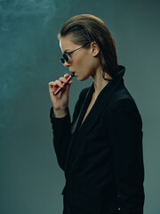 Obraz premium Woman vaping holds electronic cigarette with smoke around, wearing black suit and sunglasses in moody atmosphere, indoor setting with dark background and stylish look.