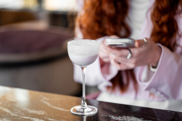 Young woman with long red hair holding smartphone at table with frothy drink in elegant glass, indoor setting, casual lifestyle moment with blurred background.