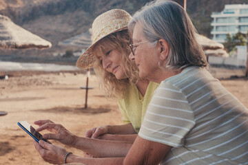 Relaxed couple of women using smartphone at the beach share social media. Senior and middle aged...