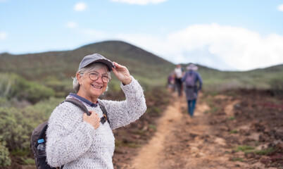 Happy senior woman with hat and eyeglasses in a trekking day in country footpath, enjoying healthy...