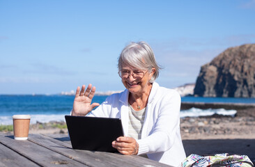 Smiling gray haired senior woman sitting relaxed near the beach using digital tablet in video chat enjoying tech and social - retired lifestyle, horizon over the sea