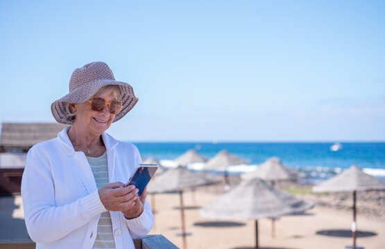 Smiling senior woman with hat and sunglasses sitting relaxed near the beach using her smartphone enjoying tech and social - retired life, horizon over the sea