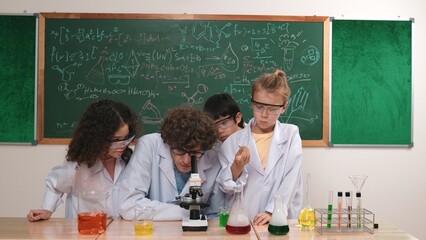 Smart caucasian teacher looking under microscope while diverse children doing experiment at STEM science lesson. Professional happy scientist inspect biochemical example by using microscope. Pedagogy.