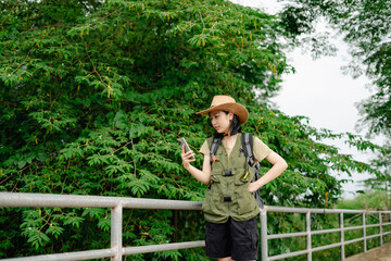 Young Asian woman in outdoor gear checks smartphone while standing next to a lush green background...