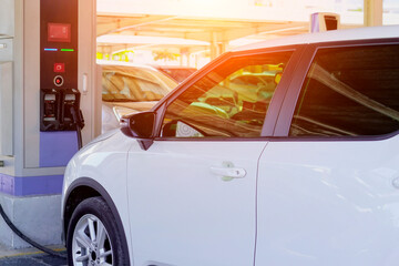 A white electric car is connected to a public charging station. The charging cable is plugged into the vehicle. Light shines on the car and station, implying a sunny day
