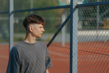 Teen Boy Stands by Fence with Serious Expression on a Sunny Court
