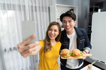 Smiling host chef influencers taking photo with smartphone by selfie together holding double big hamburger on cooking show special easy dish homemade on social media channel live streaming. Infobahn.