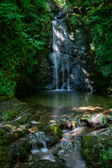 A tranquil waterfall cascades over moss-covered rocks in a lush green forest at Klaus an der...
