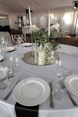Wedding reception table beautifully arranged with white roses and candles atop a mirror. Elegant tableware includes classic plates and glassware, creating a sophisticated atmosphere.