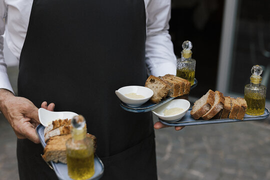 Waiter carrying rustic bread slices with olive oil and dip on modern serving trays during an outdoor event.