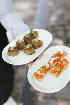 Waiter serving gourmet appetizers on white plates including bruschetta and meatballs at an outdoor event.