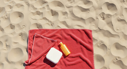 Red beach towel on sand with white sunscreen container and orange tube. Summer vacation essentials for sun protection and relaxation at seaside