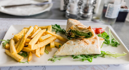 Fresh tortilla wrap served with golden fries and arugula on a white plate close-up. Casual dining, fast lunch and tasty street food in a modern restaurant setting.