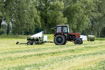 Fototapeta premium Tractor Wrapping Hay Bales with White Film in Green Meadow – Modern Agriculture in Action