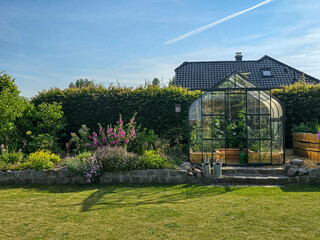 garden scene featuring a glass greenhouse, vibrant flower beds, and neatly kept lawn, all basking...