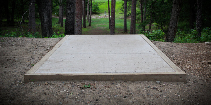 Close-up of a concrete tee pad on a disc golf course with a basket in the background. A stable and level platform made of concrete. pine tree landscape.
