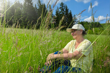 Smiling senior woman sitting on grass in nature with mountain view. Happy elderly woman with gray hair, wearing glasses and cap, sits in tall grass on meadow with forest and mountains in background