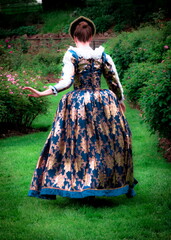 A woman in a historical dress walks through a garden.