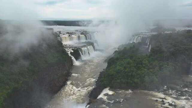 Slow motion video from the sky reveals Iguazu Falls in Brazil Multiple cascades fall into canyons as rainforest stretches beyond. Mist swirls and the drone glides over this incredible scene of nature