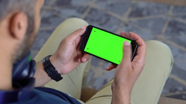 Man on sofa holding phone in horizontal position with green screen &ndash; top down view for media replacement