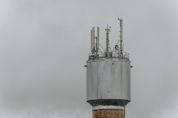 A tall communication tower with antennas rises against a cloudy sky, featuring a brick base and a metal structure at the top equipped with telecom equipment.
