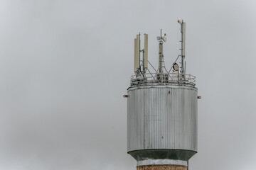A tall communication tower with antennas rises against a cloudy sky, featuring a brick base and a metal structure at the top equipped with telecom equipment.