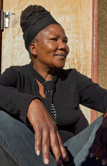 village single african woman with dreadlocks braids sited in front of the house , in the yard, wearing headwrap with a smile