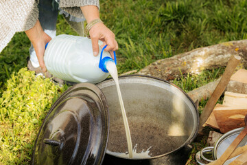 Fresh milk is being poured from a plastic container into a large metal pot outdoors, surrounded by...