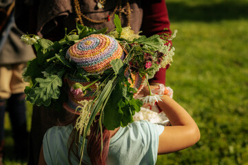 A child with a flower-adorned crochet hat watches a large pot over an open fire, suggesting...