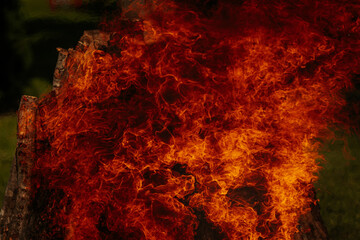 Tall bonfire burning intensely with orange flames reaching high, surrounded by rocks and trees...