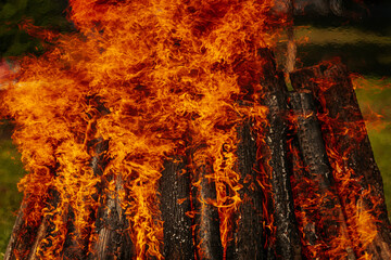 Tall bonfire burning intensely with orange flames reaching high, surrounded by rocks and trees...