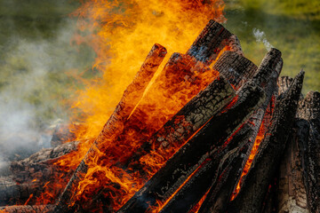 Intense orange flames engulf burning firewood in a large bonfire, with vivid textures of charred...