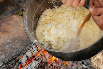 Traditional cheese curds with caraway seeds cook in a pot over open fire, stirred with a wooden...